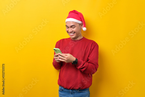 Cheerful young Asian man in Santa hat using smartphone for messaging with friends over yellow studio background. celebration Christmas holiday and New Year concept