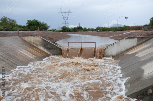 Rainwater runoff flows through a concrete stormwater drainage canal, featuring a central grate and turbulent muddy water under an overcast sky with vegetation and power lines nearby