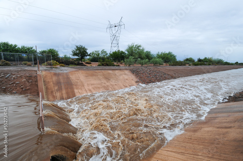 A rainwater runoff canal channeling muddy stormwater through a concrete structure; the rushing water contrasts with the desert vegetation and overcast sky, showcasing effective drainage design