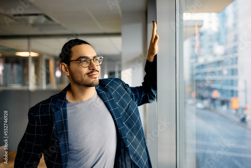 Smiling Hispanic entrepreneur day dreaming while looking through window of his office.