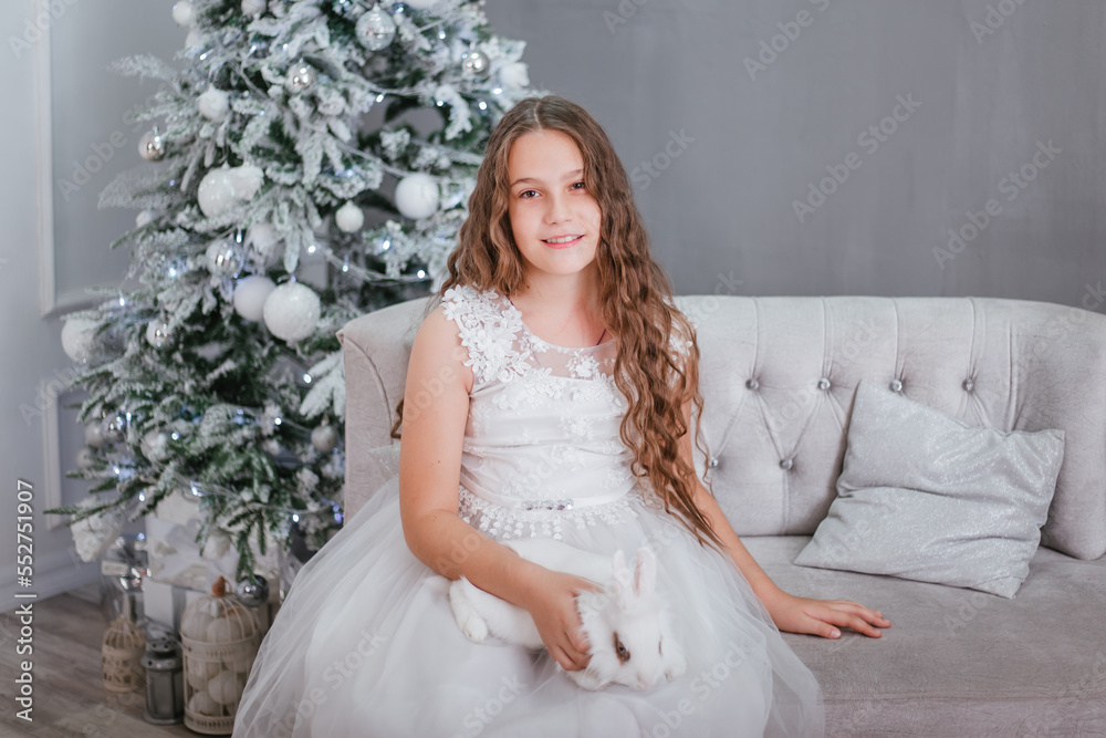 A cute girl with long wavy hair in a white dress on the background of a Christmas tree with a rabbit in her hands. A cute girl with long wavy hair in a white dress on the background of a Christmas tree with a rabbit in her hands.
