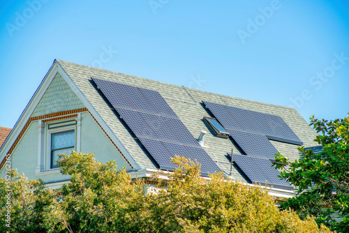 Modern house facade with large solar pannels on roof with light tiles and green stucco with front yard trees and foliage in neighborhood © Aaron
