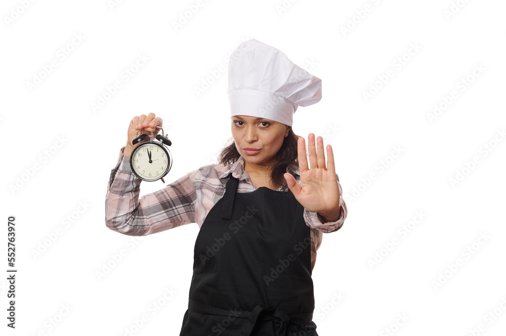 Serious multi-ethnic woman waitress, cafe worker in black apron holding black alarm clock showing opening and closing time, extending her hand to show stop or prohibition gesture, on white background. Serious multi-ethnic woman waitress, cafe worker in black apron holding black alarm clock showing opening and closing time, extending her hand to show stop or prohibition gesture, on white background.