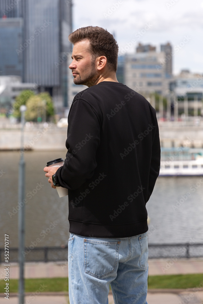 City portrait of handsome hipster guy with beard wearing black blank hoodie or sweatshirt with space for your logo or design. Holding coffee in his hands. Mockup for print