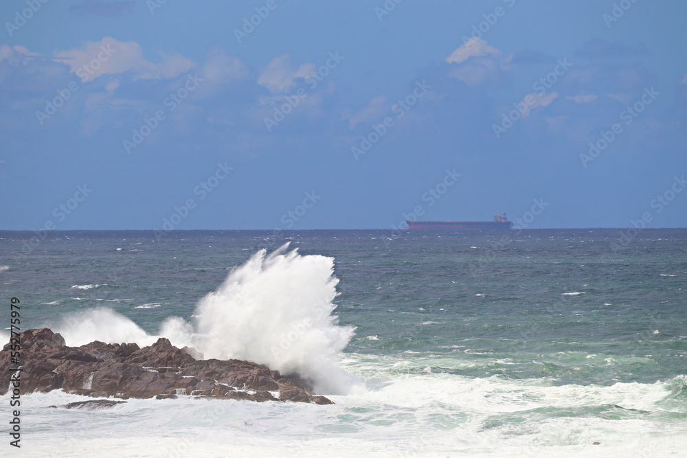 Waves crashing over the rocks near Fingal Bay, with a container or cargo ship in the background.
