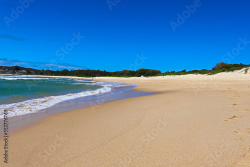 Waves crashing at Fingal Beach with people walking along the sand in the distance on a sunny day.