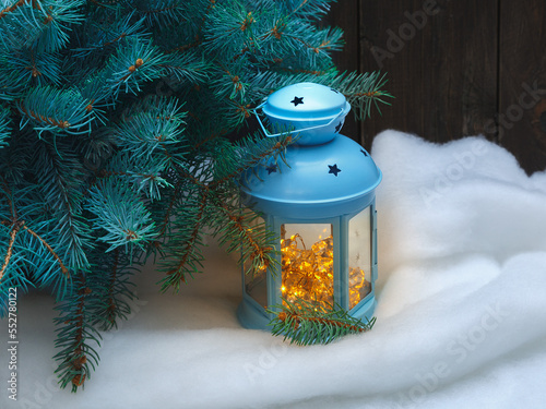A Christmas lantern with a luminous garland inside stands under a Christmas tree on the snow