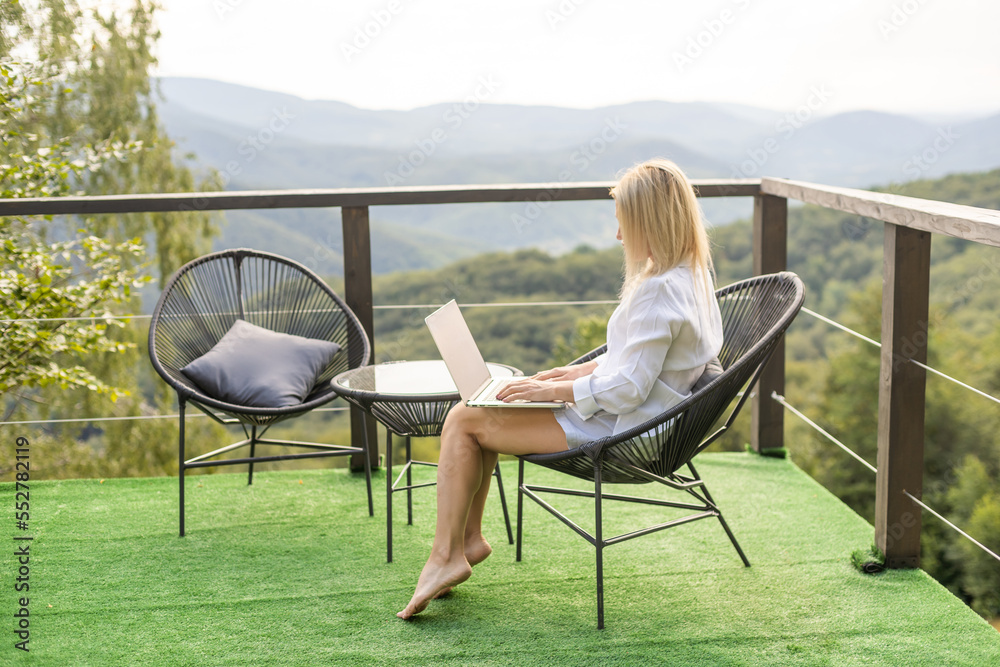 A young woman, freelancer is working on a laptop remotely on a balcony in the open fresh air near the mountains in warm sunny weather.