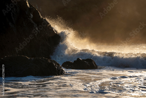 Majestic landscape image of jade turquoise waves crashing onto shore and rocks in Kynance Cove Cornwall with glowing sunrise background and water spray dorplets in wind