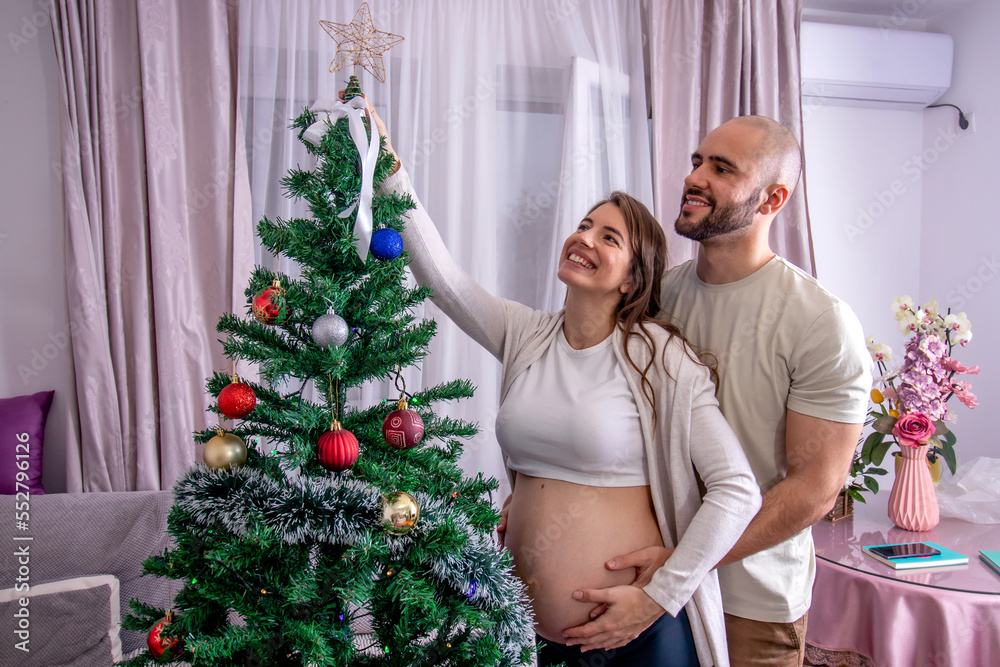 Pregnant woman with her partner decorating the Christmas tree. A young loving couple preparing for childbirth. Best Christmas gift.