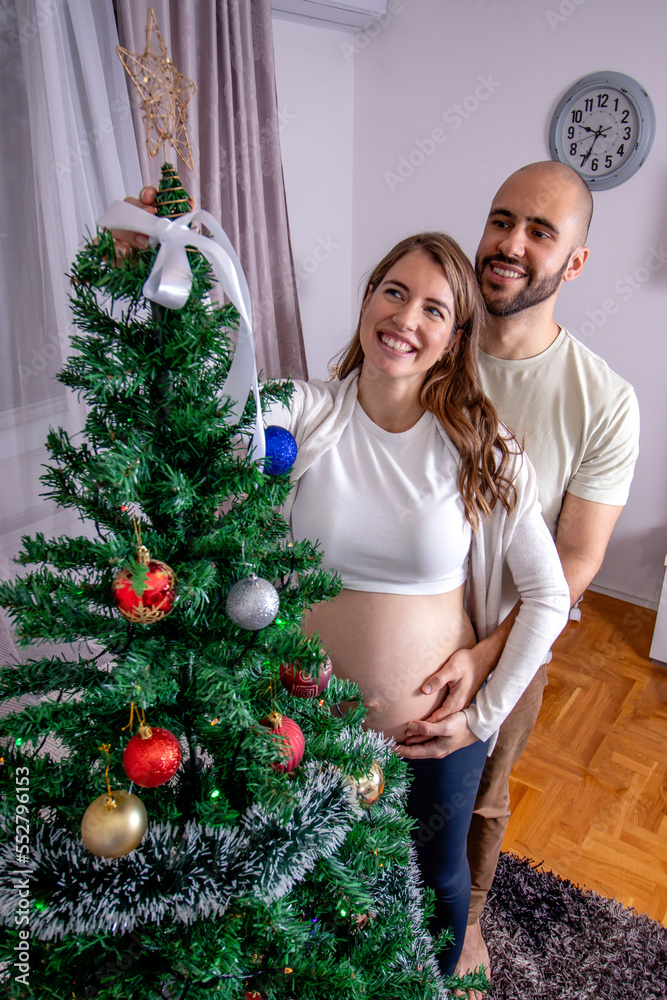 Pregnant woman with her partner decorating the Christmas tree. A young loving couple preparing for childbirth. Best Christmas gift.