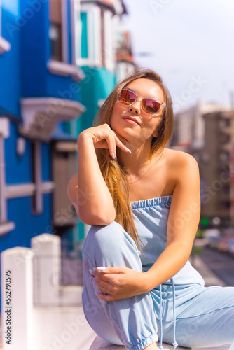 Fototapeta Naklejka Na Ścianę i Meble -  Portrait of attractive young blonde woman wearing sunglasses, behind blue colorful facade