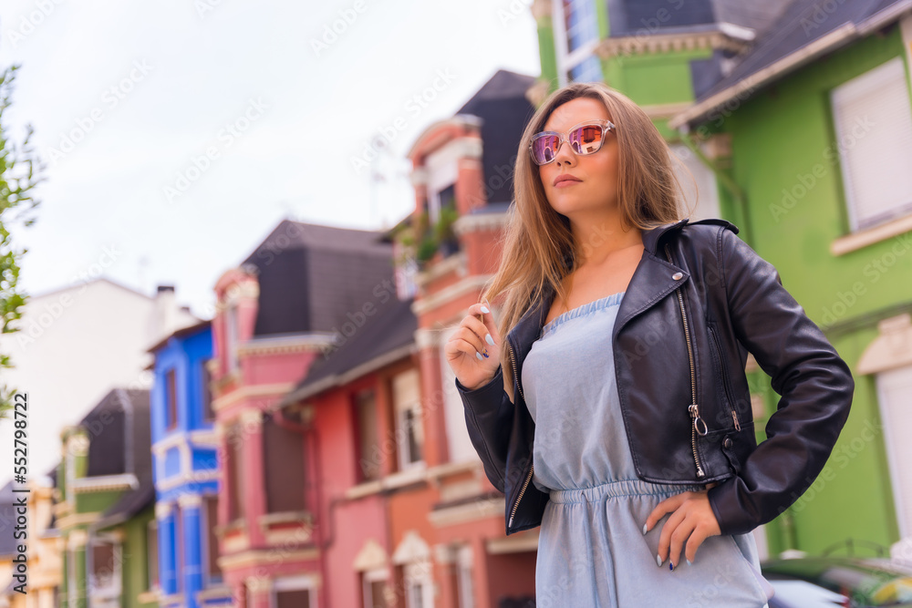 Attractive young blonde woman in leather jacket and sunglasses, behind colorful facade