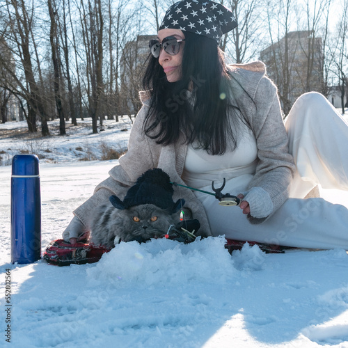 Winter fishing with a cat. A funny British fluffy cat in mittens and a knitted hat and a girl in sunglasses and a bandana on her head are fishing with winter fishing rods on the ice of the lake.