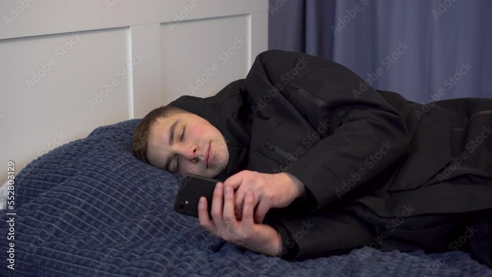 A young man in a jacket lies on the bed and uses a smartphone. The man did not pay the bills and was left without heating in the winter. A young man in a jacket lies on the bed and uses a smartphone. The man did not pay the bills and was left without heating in the winter.