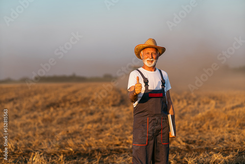Senior man is standing in a field being plowed, he is holding a digital tablet in his hand and showing a thumbs up.