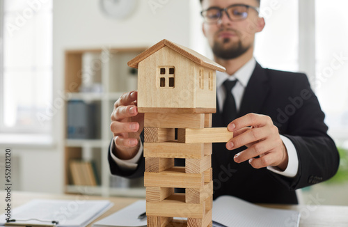 Close up of little miniature wooden toy house with tower foundation where young entrepreneur removes blocks as symbol of business risks and risky dangerous situation on real estate construction market