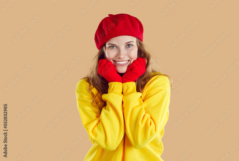 Portrait of cute smiling young woman in autumn clothes holding her hands near her face. Caucasian girl in red beret, gloves and yellow sweatshirt is smiling on light beige background. Close up. Portrait of cute smiling young woman in autumn clothes holding her hands near her face. Caucasian girl in red beret, gloves and yellow sweatshirt is smiling on light beige background. Close up.