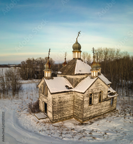 Dilapidated orthodox church set in snow covered farmland with thin white clouds in a blue sky. 