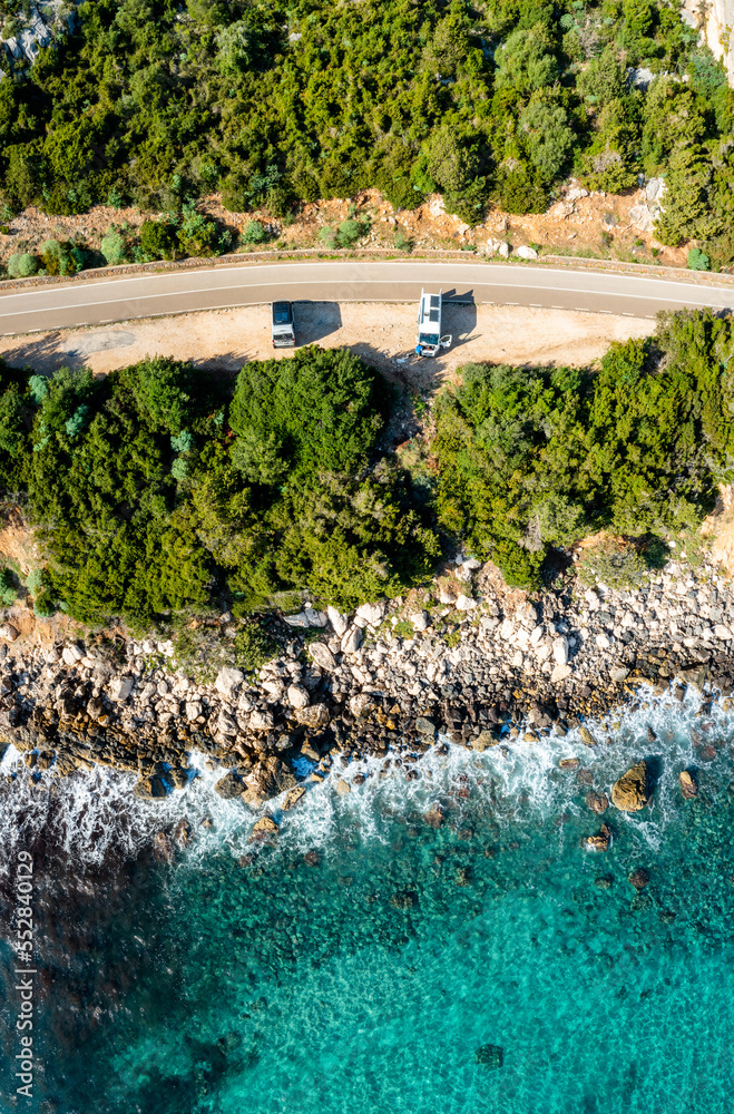 top down view of two camper vans parked on a narrow coastal road in the forest with turquoise water and rocky shore
