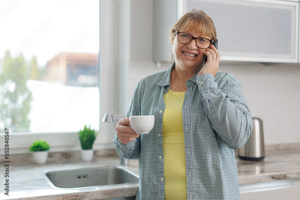 Shot of smiling adult woman using her mobile phone while drinking a cup of coffee in the kitchen at home. Shot of smiling adult woman using her mobile phone while drinking a cup of coffee in the kitchen at home.
