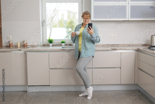 Shot of smiling adult woman using her mobile phone while drinking a cup of coffee in the kitchen at home.