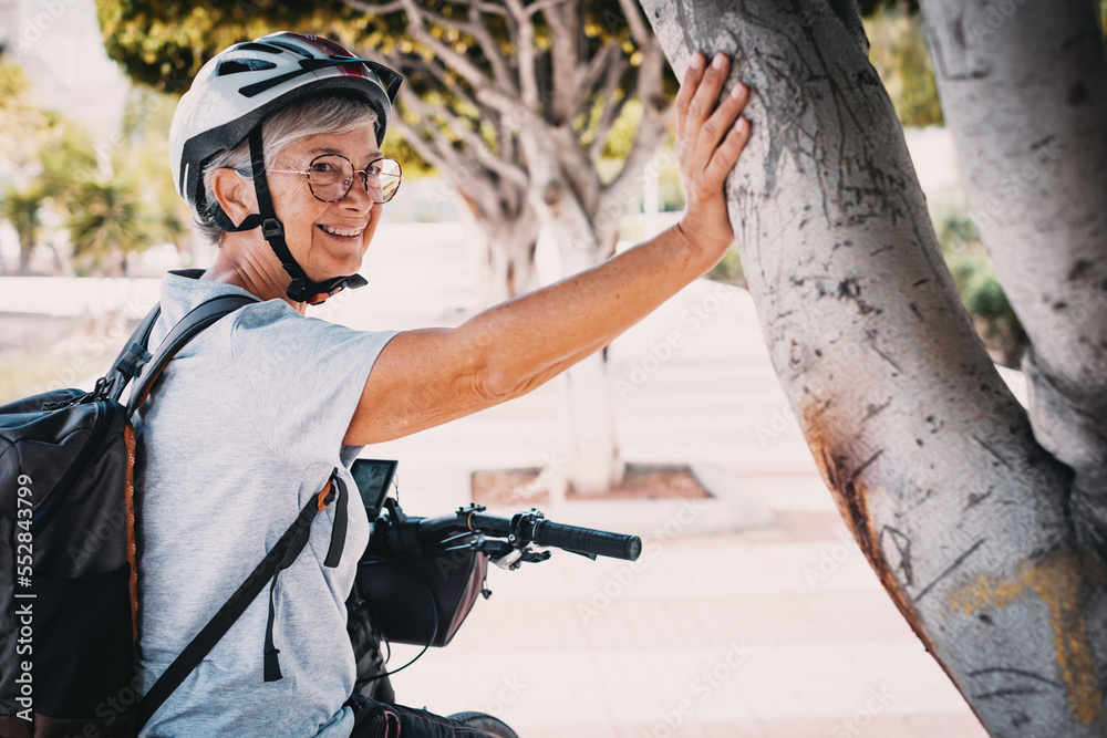 Fototapeta premium Cheerful cyclist elderly woman in urban park wearing helmet and backpack running with her electro bicycle. Concept of healthy lifestyle and sustainable mobility
