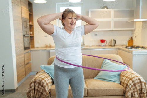 Overweight mature woman in white t-shirt and gray leggings trying to lose weight with hula hoop exercises photo