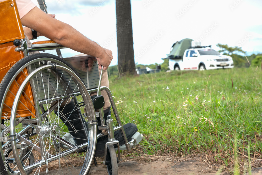 Naklejka premium Closeup of man' s hand sitiing on wheechair, witt blur background of pickup truck camping tent on tawn of camping ground, to people wheelchair and tourists concept