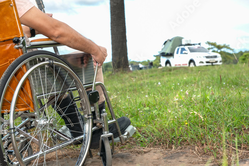 Closeup of man' s hand sitiing on wheechair, witt blur background of pickup truck camping tent on tawn of camping ground, to people wheelchair and tourists concept