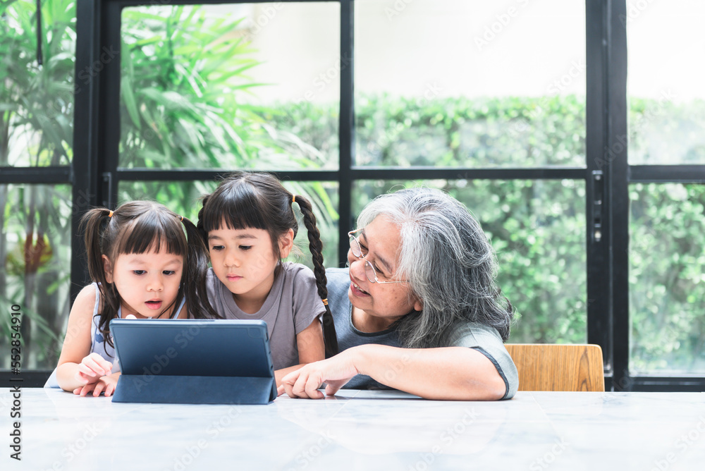 Asian family, Elderly grandmother and 2 daughters looking at tablet on marble table in the house, with blur background, to Asian family and children education online concept. Asian family, Elderly grandmother and 2 daughters looking at tablet on marble table in the house, with blur background, to Asian family and children education online concept.