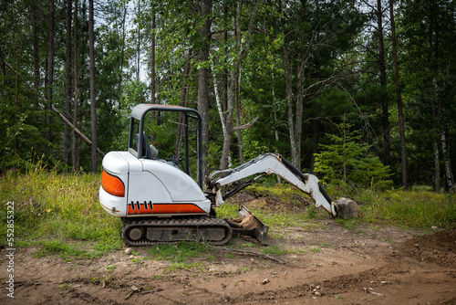 Mini excavator machine with extended arm, at a new home construction site, near exposed dirt or soil and forest trees.