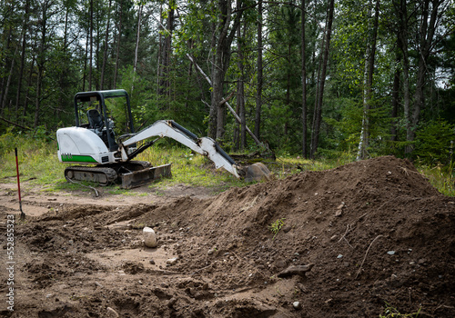 Mini excavator machine with extended arm, at a new home construction site, behind piles of exposed dirt or soil and near forest trees.