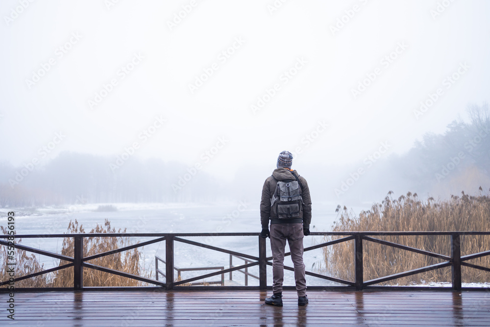 Young man in fog standing alone on wooden footbridge and staring at lake. Hooded guy. Peaceful atmosphere in nature. Enjoying fresh air in winter. Back view.