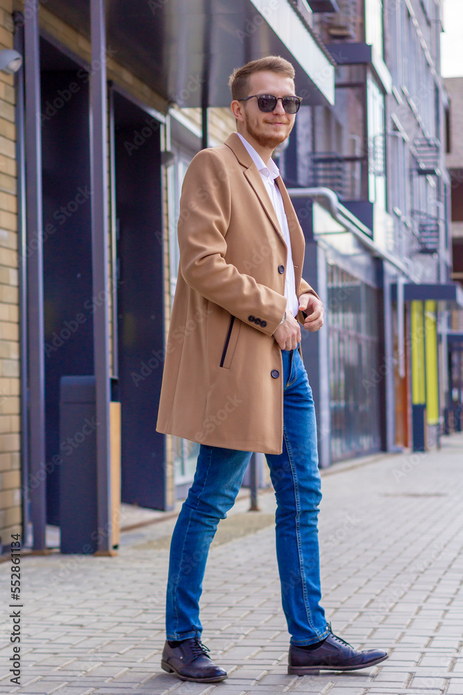 A young man dressed in a beige coat is walking down the street. A young bearded guy with a modern hairstyle and sunglasses on an urban background