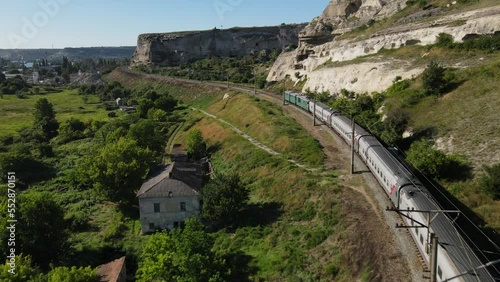 A cinematic flight behind a train traveling near a white cliff. Single railway. A green locomotive with grey carriages. A river flowing into the sea. Monastery in the rock.