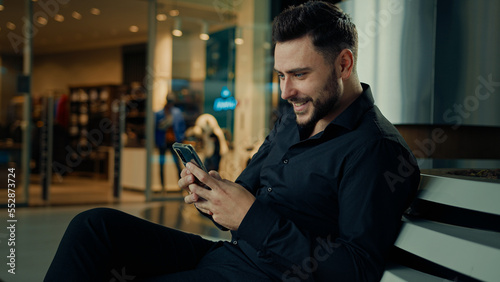 Caucasian stylish man in black suit successful businessman customer sit at shopping mall using mobile phone apps make online order buying clothes win internet discounts chatting distant communication