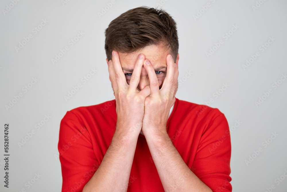 Scared man hides face behind palms, peeks through fingers, feels frightened Scared man hides face behind palms, peeks through fingers, feels frightened