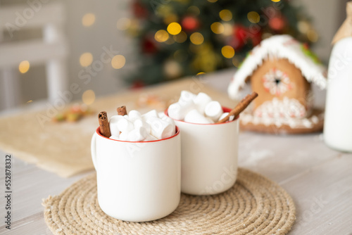 two white cup of cocoa with marshmallows and gingerbread house on table on background of Christmas tree