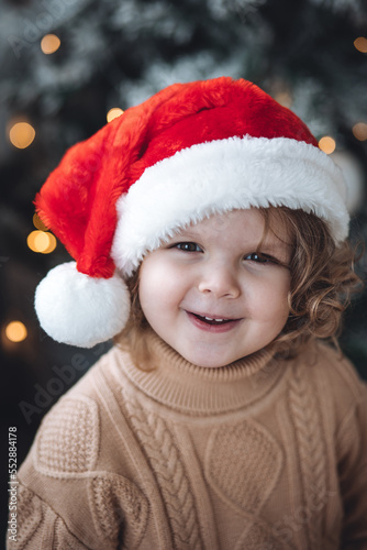 Portrait of a cute little girl in a beautiful knitted sweater ad a red Santa hat near decorated Christmas and new year tree at home. Smiling, cheerful and happy