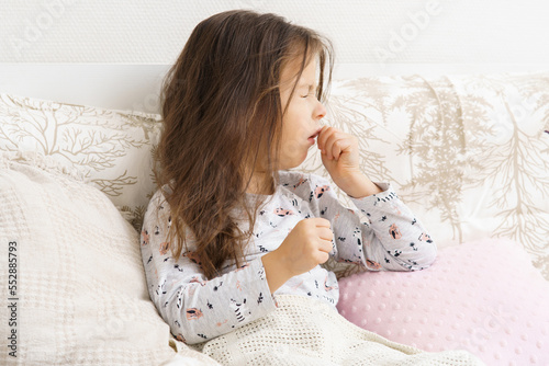 Poor, unfortunate little brunette preschooler child, girl in pajamas coughing, lying in soft bed. Sore throat, angina, tonsillitis. Recovering, health care, home treatment and bed rest. Copy space