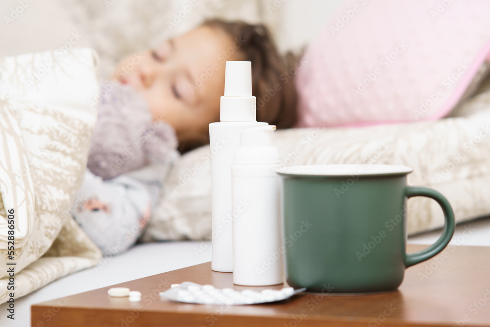 Close up green mug, nasal and throat sprays containers and pills blister on bed table against defocused sick little child, girl sleeping in bed. Pharmacy and remedy as painkillers, antibiotics. Close up green mug, nasal and throat sprays containers and pills blister on bed table against defocused sick little child, girl sleeping in bed. Pharmacy and remedy as painkillers, antibiotics.