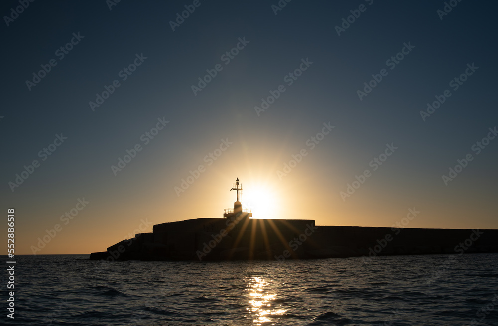 The sun goes down on the horizon. A wall by the sea with a lighthouse stands in front of it as a contrast. The sky and the water are blue.