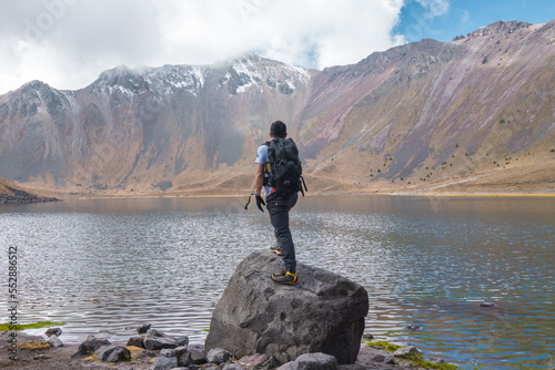 horizontal shot of man with backpack looking at a lake with snowy mountains on a sunny day