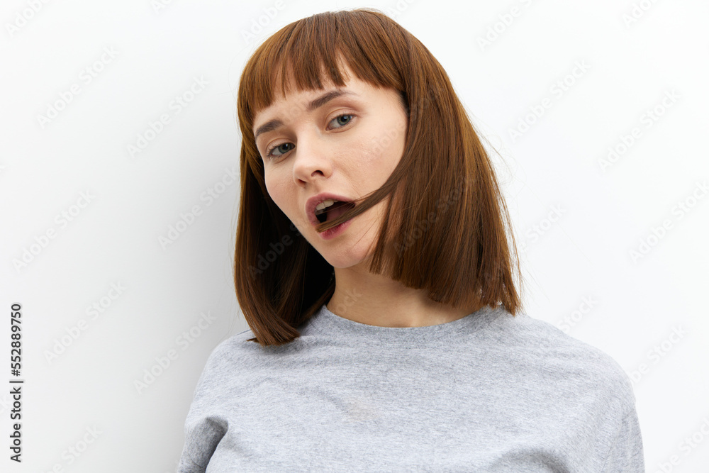 a beautiful, attractive woman stands in a grey T-shirt on a light background and playfully bites a lock of her red hair a beautiful, attractive woman stands in a grey T-shirt on a light background and playfully bites a lock of her red hair