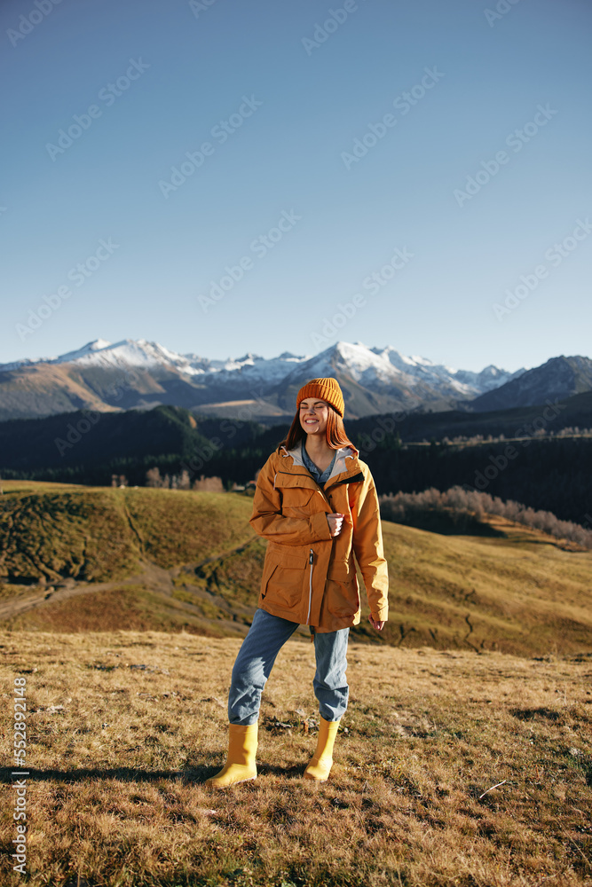 Autumn woman walking up the hill in full smile travels to the mountains in nature hiking and happiness in a yellow cape against the snowy mountains in the sunset, freedom of life style