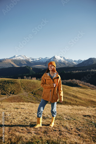 Autumn woman walking up the hill in full smile travels to the mountains in nature hiking and happiness in a yellow cape against the snowy mountains in the sunset, freedom of life style