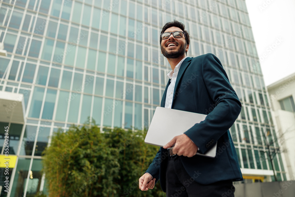 Smiling indian businessman in suit and glasses with laptop near office building Smiling indian businessman in suit and glasses with laptop near office building