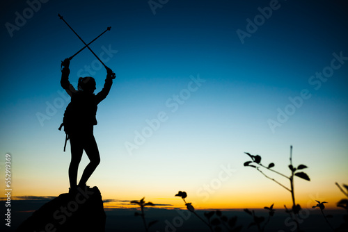 Silhouette of a hiker girl on a rock pedestal with hands up. Beautiful orange sunset on blue sky. Tree branches with leaves in the foreground.