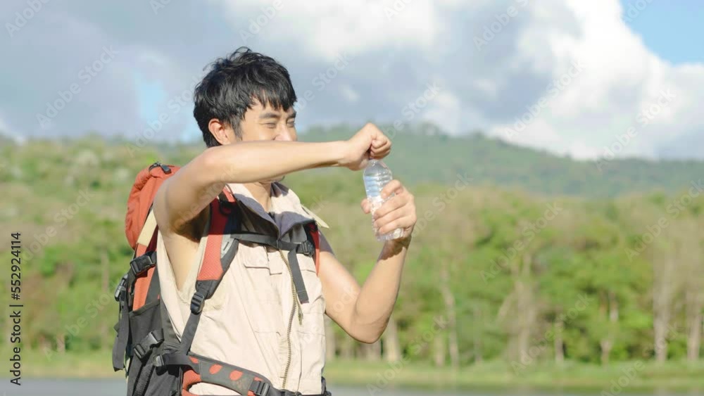 Young Asian man backpacker traveling in the forest. Male traveler holds a bottle of water. Healthy hiker drinking water in nature hike. Young man living active life, fun and smile.